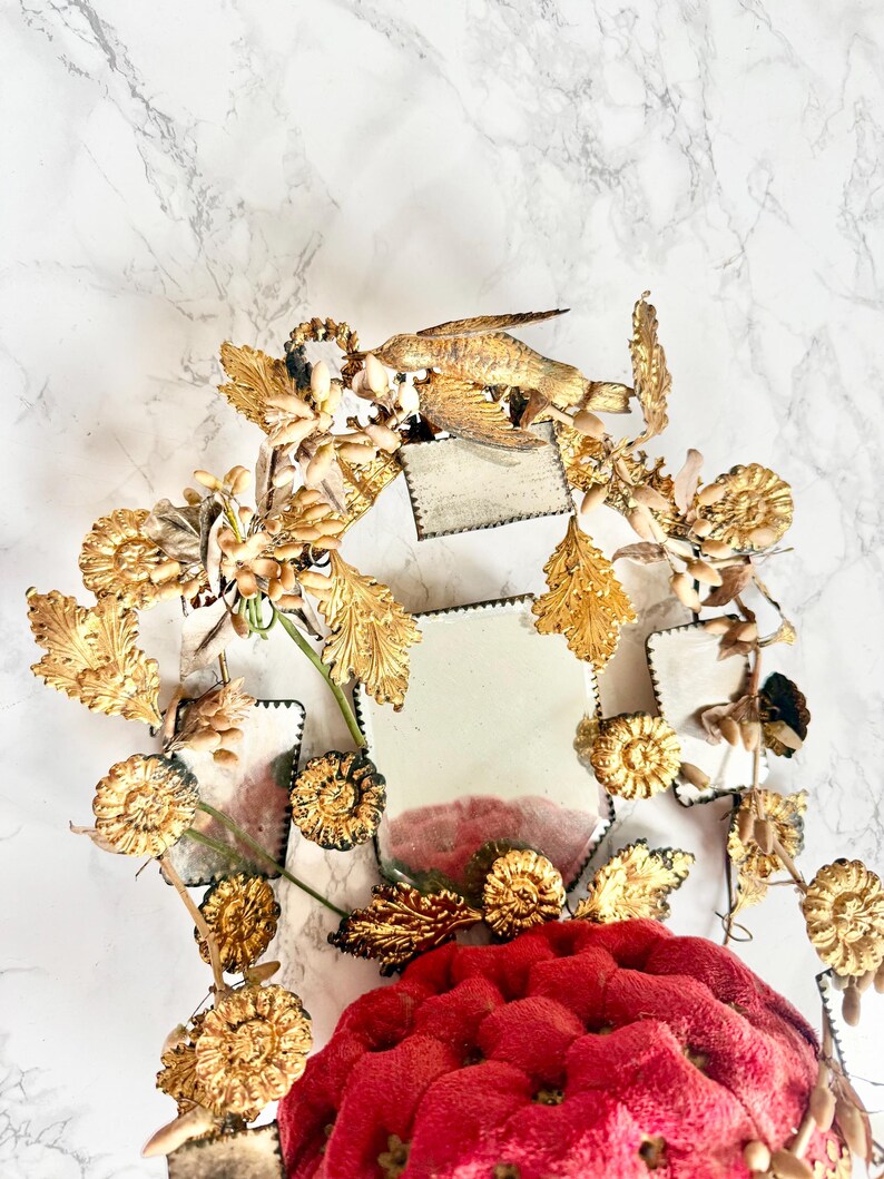 May include: A decorative mirror with a gold-coloured frame and a red, tufted cushion base. The frame is adorned with detailed floral and bird-like elements. The mirror's surface is divided into several small, square sections.