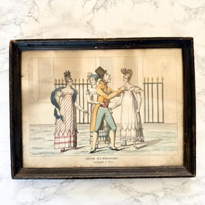 Peut inclure: Une estampe ancienne encadrée représentant une scène avec des personnages en costume d'époque. Le cadre en bois noir entoure une estampe montrant un homme en manteau jaune et chapeau haut-de-forme avec deux femmes. Le texte "LEÇON AUX ETRANGERS" est visible.