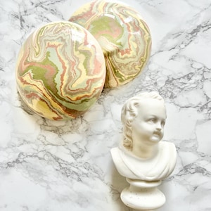 May include: Two round, decorative bowls with a marbled design in shades of green, yellow, and pink. The bowls are sitting on a white marble surface. A white ceramic bust of a child is also on the surface.