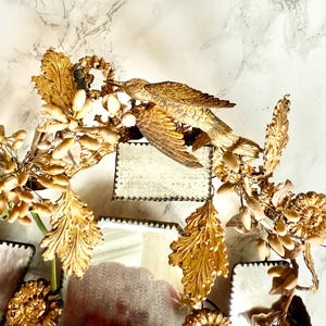 May include: A decorative mirror with a gold finish, featuring a bird and floral motif. The mirror has a distressed appearance and is adorned with detailed, gilded leaves and a bird figurine. The design has a vintage and elegant feel.