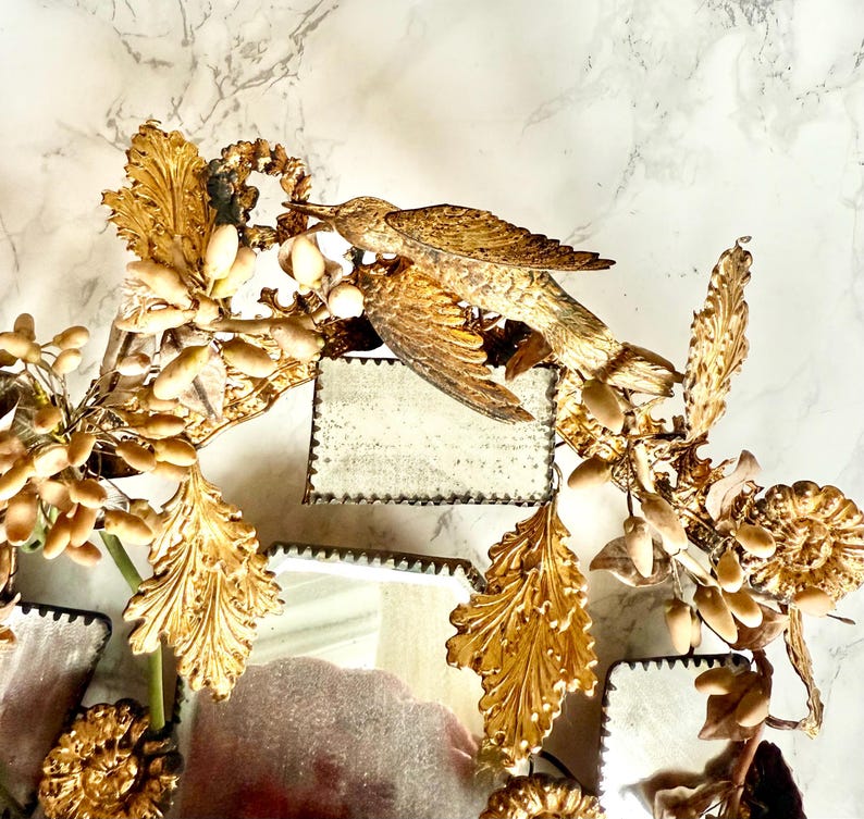 May include: A decorative mirror with a gold finish, featuring a bird and floral motif. The mirror has a distressed appearance and is adorned with detailed, gilded leaves and a bird figurine. The design has a vintage and elegant feel.