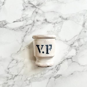 Puede incluir: Jarrón de cerámica blanca con una inscripción azul y negra que dice "V.F."