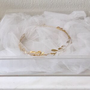 May include: A gold leaf crown sits on a bed of white tulle in a clear acrylic box.