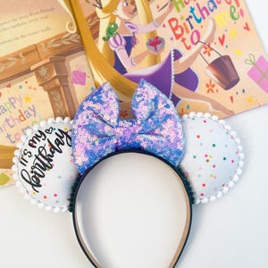 May include: A black headband with white ears and a large sequined bow. The ears are decorated with confetti and pom-poms. The bow is blue and purple. The text "It's my birthday" is on one ear. A children's book is in the background.