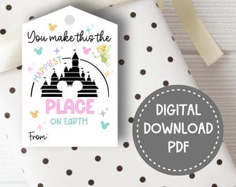 May include: A white rectangular gift tag with a hole at the top, featuring a black silhouette of a castle, a fairy illustration, and the text "You make this the HAPPIEST PLACE ON EARTH" in black, pink, and light blue, with a "From:" line at the bottom.