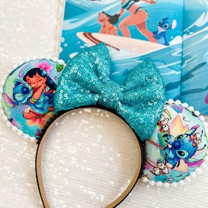 May include: A pair of mouse ears featuring a large, turquoise sequined bow. The ears are decorated with colorful images of Lilo & Stitch characters. The headband is black with a tan interior. A children's book with a surfing scene is in the background.