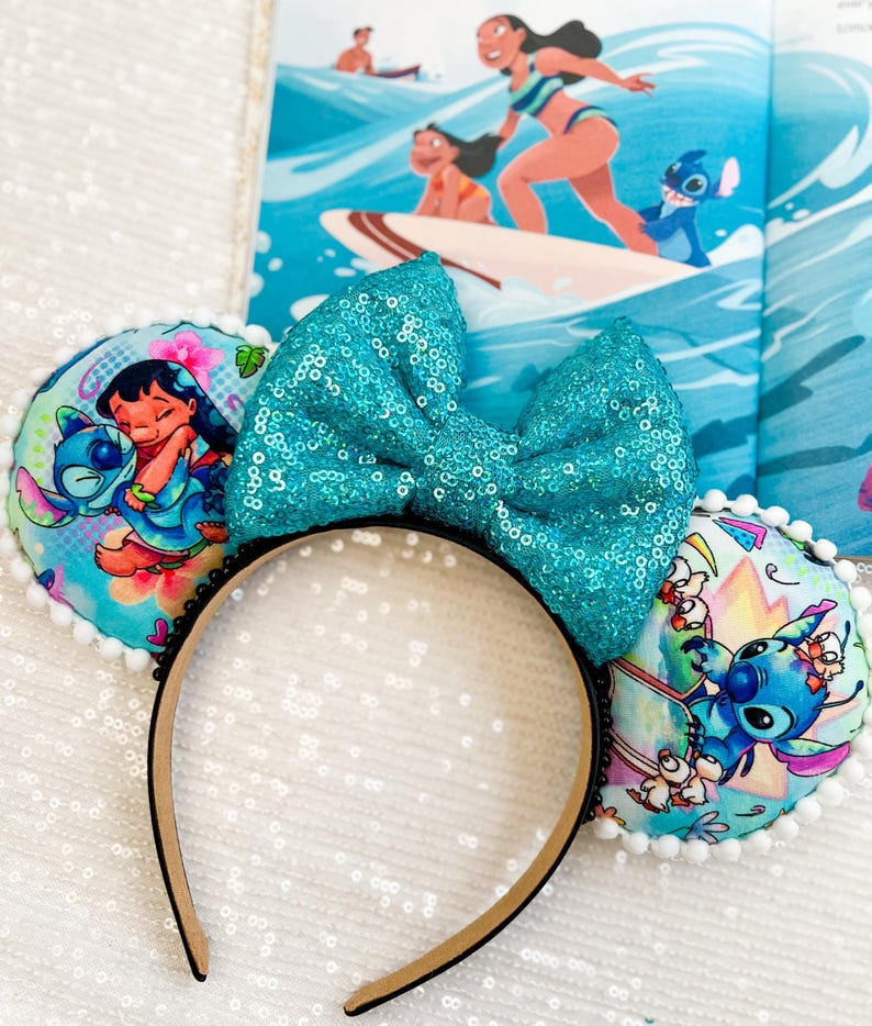 May include: A pair of mouse ears featuring a large, turquoise sequined bow. The ears are decorated with colorful images of Lilo & Stitch characters. The headband is black with a tan interior. A children's book with a surfing scene is in the background.