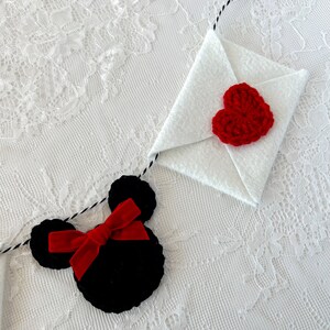 May include: A decorative garland featuring a black crocheted mouse head shape with a red velvet bow and a white felt envelope adorned with a red crocheted heart, all strung together on a black and white twisted cord for hanging display.