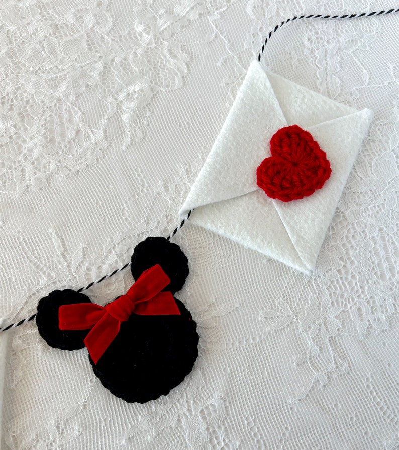 May include: A decorative garland featuring a black crocheted mouse head shape with a red velvet bow and a white felt envelope adorned with a red crocheted heart, all strung together on a black and white twisted cord for hanging display.