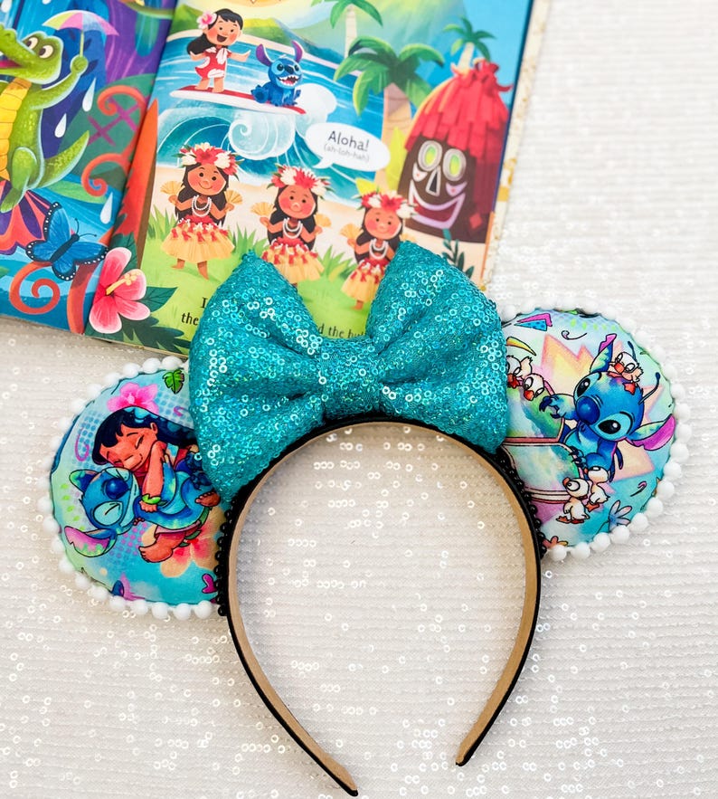 May include: A teal sequined bow headband with round ear accents featuring Lilo & Stitch characters. The headband has a black band and a light-colored interior. The ears have a white pom-pom trim. A children's book with illustrations is in the background.
