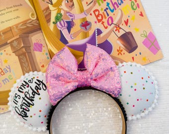 May include: A pink sequined bow headband with black trim and a tan interior. The headband has two white circular ears with colorful confetti and a pom-pom trim. A children's book with a Rapunzel illustration and the text "Happy Birthday to me!" is in the background.