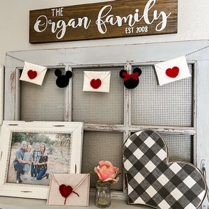 May include: A decorative garland featuring white felt envelopes with red crocheted hearts and black mouse-shaped silhouettes, one with a red bow. Below, a framed family photo, a tan envelope with a red heart, a pink flower, and a buffalo check heart.