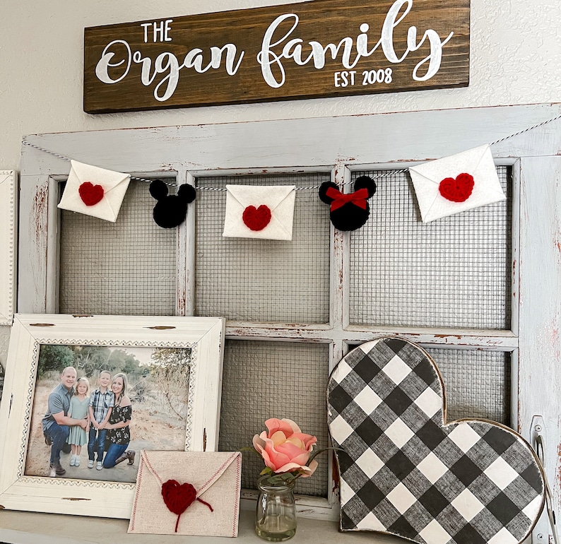 May include: A decorative garland featuring white felt envelopes with red crocheted hearts and black mouse-shaped silhouettes, one with a red bow. Below, a framed family photo, a tan envelope with a red heart, a pink flower, and a buffalo check heart.