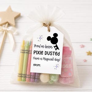 May include: A sheer white organza drawstring bag containing colorful crayons, small round candies, and a pink bubble bottle. A white rectangular tag attached with twine features a black silhouette of a key with mouse ears and the text You've been PIXIE DUSTED Have a Magical day! FROM: