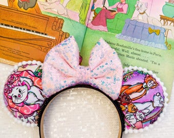 May include: A black headband with a pink sequined bow and two round ears featuring illustrations of cats. The ears have white pom-pom trim. The background includes an open book with illustrations of cats and text.