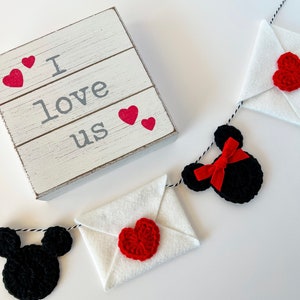 May include: A white wooden sign with the text I love us and four small red hearts, accompanied by a garland featuring black crocheted mouse-shaped silhouettes, white felt envelopes, red crocheted hearts, and a red ribbon bow on a black and white string.