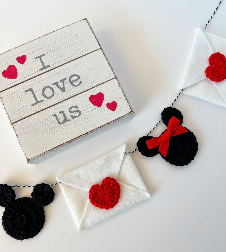 May include: A white wooden sign with the text I love us and four small red hearts, accompanied by a garland featuring black crocheted mouse-shaped silhouettes, white felt envelopes, red crocheted hearts, and a red ribbon bow on a black and white string.