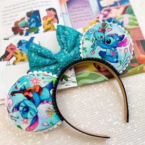May include: A headband featuring two round ears with Stitch illustrations and a large, teal sequined bow. The ears have a white scalloped trim and are decorated with Stitch playing a guitar and hugging a doll. The headband is black with black bead accents.