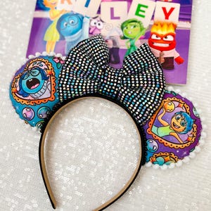 May include: A black headband with Inside Out themed ears. The ears feature images of characters and a sequined bow. The word "RILEY" is visible in the background. The ears are trimmed with white pom-poms.