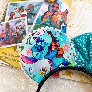 May include: A blue and pink Stitch character ear headband. The headband features a cartoon Stitch playing a guitar against a light blue floral background. The headband is trimmed with white pom-poms and a black beaded band.