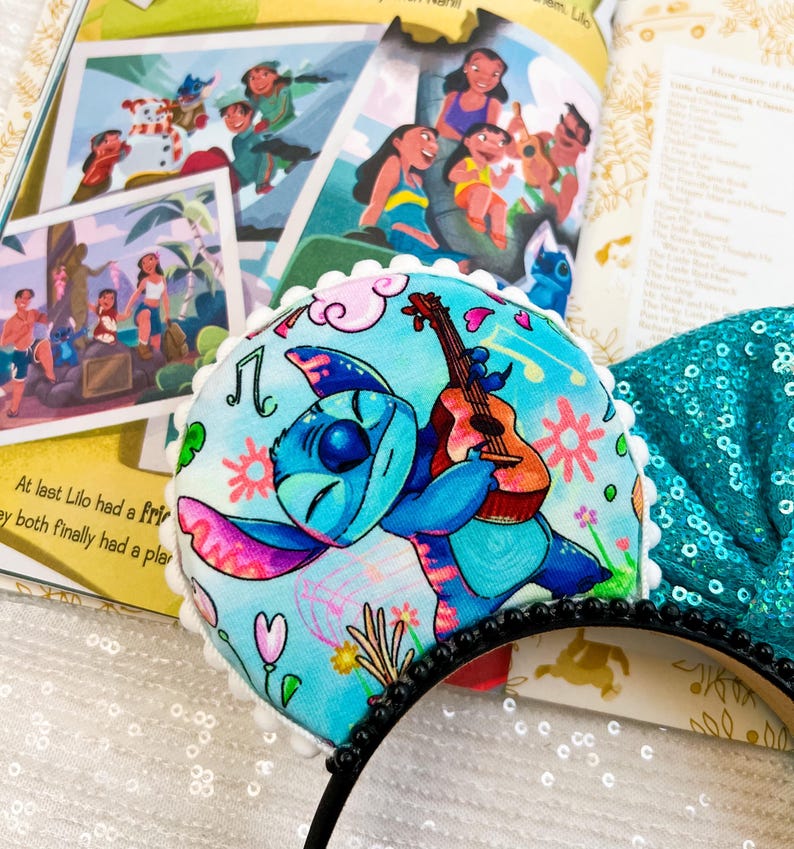 May include: A blue and pink Stitch character ear headband. The headband features a cartoon Stitch playing a guitar against a light blue floral background. The headband is trimmed with white pom-poms and a black beaded band.