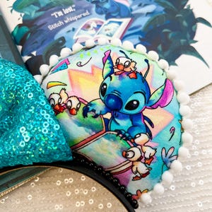 May include: A pair of black mouse ears with a Stitch design. The ears feature a colorful print of Stitch and ducklings, trimmed with white pom-poms. A teal sequined bow is visible. The book behind the ears has the text "I'm lost." Stitch whispered.