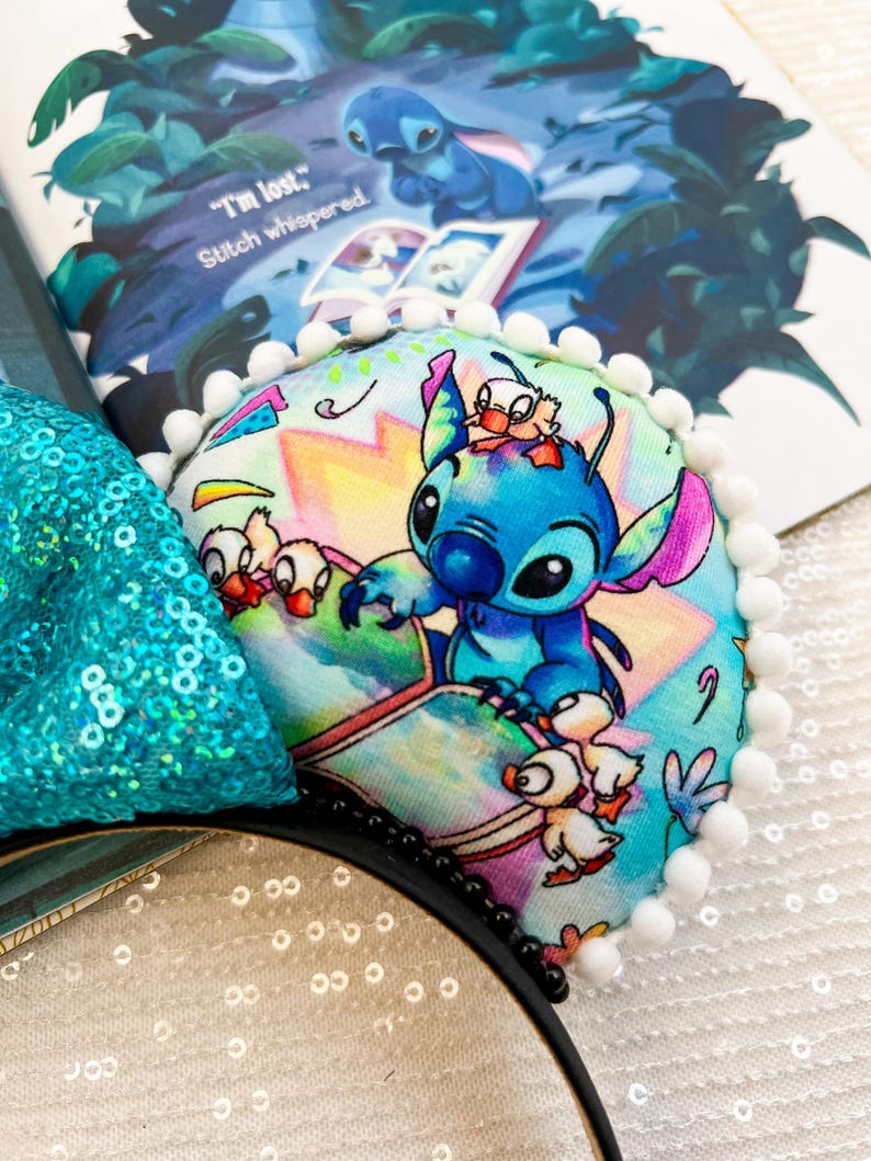 May include: A pair of black mouse ears with a Stitch design. The ears feature a colorful print of Stitch and ducklings, trimmed with white pom-poms. A teal sequined bow is visible. The book behind the ears has the text "I'm lost." Stitch whispered.