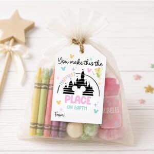 Pode incluir: Uma sacola de presente de celofane transparente amarrada com barbante, contendo giz de cera, doces e um frasco de bolhas rosa. Uma etiqueta diz "You make this the happiest place on Earth" com um gr&aacute;fico de castelo. Uma varinha de estrela de madeira est&aacute; no fundo.