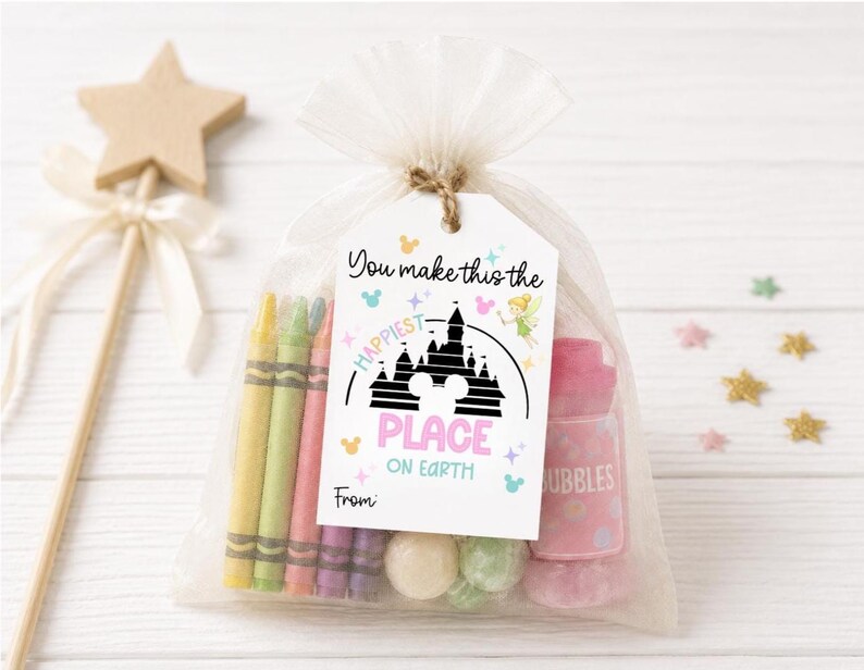 Pode incluir: Uma sacola de presente de celofane transparente amarrada com barbante, contendo giz de cera, doces e um frasco de bolhas rosa. Uma etiqueta diz "You make this the happiest place on Earth" com um gr&aacute;fico de castelo. Uma varinha de estrela de madeira est&aacute; no fundo.