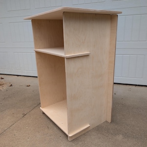 May include: A light-colored, unfinished wooden cabinet with two shelves and a top surface. The cabinet is rectangular with a simple, modern design. The wood grain is visible, and the cabinet appears to be made of plywood.