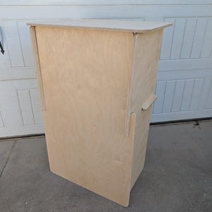 May include: A tall, rectangular stand constructed from light-colored plywood. The structure features a flat top surface and vertical side panels with visible interlocking joints, creating a clean, functional, and upright display unit.