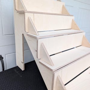 May include: A wooden display shelf with a stepped design. The shelf is made of light-colored plywood and features three steps, supported by triangular brackets. The shelf is designed to be placed against a wall.