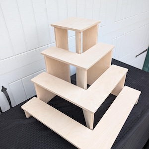 Puede incluir: Un conjunto de cuatro elevadores de exhibición de madera sin terminar en una configuración escalonada. Los elevadores son de madera de color claro y están dispuestos en un diseño en forma de L escalonado. El elevador más grande está en la parte inferior.