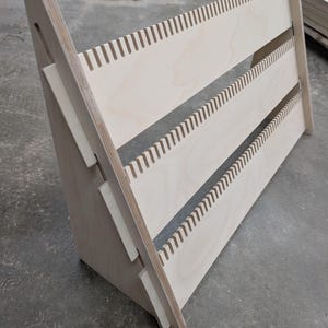 Puede incluir: Una estantería de madera clara con tres estantes. Los estantes tienen una serie de ranuras verticales espaciadas uniformemente. Los soportes laterales están en ángulo, creando una forma triangular. La estantería es de madera sin terminar.