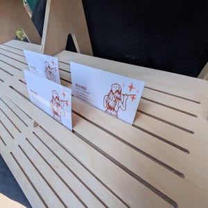 K&ouml;nnte beinhalten: Ein heller, mehrstufiger Pr&auml;sentationsst&auml;nder aus Holz mit mehreren horizontalen Schlitzen f&uuml;r Karten. Zwei wei&szlig;e Visitenkarten mit einer roten Strichzeichnung einer Person und dem Text Kelci Buss Owner & Designer stecken in den Schlitzen.
