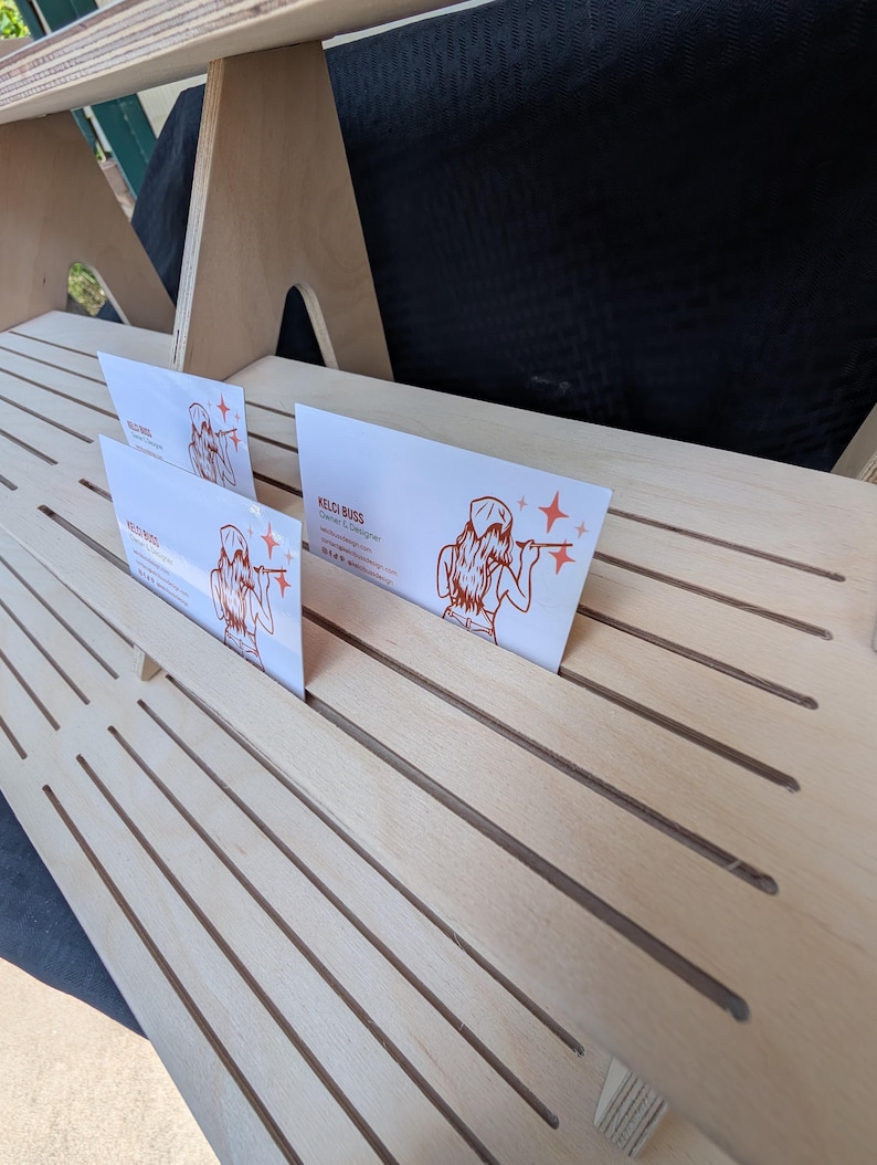 K&ouml;nnte beinhalten: Ein heller, mehrstufiger Pr&auml;sentationsst&auml;nder aus Holz mit mehreren horizontalen Schlitzen f&uuml;r Karten. Zwei wei&szlig;e Visitenkarten mit einer roten Strichzeichnung einer Person und dem Text Kelci Buss Owner & Designer stecken in den Schlitzen.