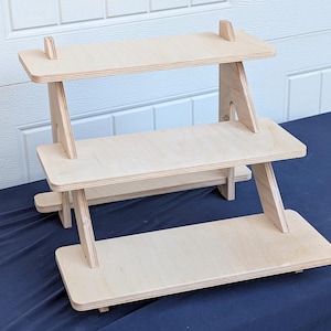 May include: Two-tiered wooden display stand with a natural finish. The stand has a rectangular base and two shelves, each supported by angled legs. The top shelf has two small wooden supports. The stand is designed for displaying items.
