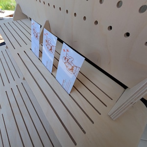 K&ouml;nnte beinhalten: Ein mehrstufiger Pr&auml;sentationsst&auml;nder aus hellem Holz mit horizontalen Schlitzen und einer R&uuml;ckwand mit Reihen von kreisf&ouml;rmigen L&ouml;chern. Drei rechteckige wei&szlig;e Visitenkarten mit orangefarbenen Strichzeichnungen sind in die Schlitze gesteckt.