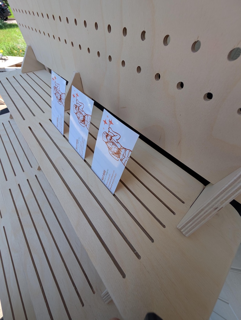 K&ouml;nnte beinhalten: Ein mehrstufiger Pr&auml;sentationsst&auml;nder aus hellem Holz mit horizontalen Schlitzen und einer R&uuml;ckwand mit Reihen von kreisf&ouml;rmigen L&ouml;chern. Drei rechteckige wei&szlig;e Visitenkarten mit orangefarbenen Strichzeichnungen sind in die Schlitze gesteckt.