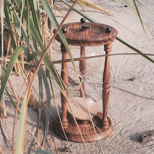 Op de afbeelding: Een houten zandloper met een met zand gevulde glazen bol, geplaatst op zand. De zandloper heeft een gedraaid houten frame met een donkere afwerking. Het zand stroomt van de bovenste bol naar beneden. De scène wordt omlijst door grassprieten.