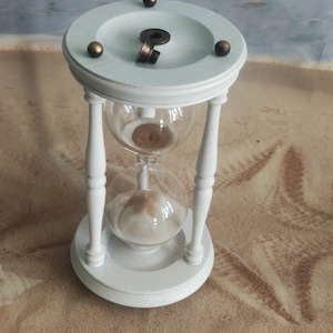 May include: White hourglass with a wooden base and brass accents. The hourglass is filled with sand and is sitting on a sandy surface.