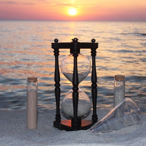 Può includere: Una clessidra nera con la sabbia che scorre attraverso il bulbo superiore, seduta su una spiaggia sabbiosa con due bottiglie di vetro trasparente contenenti sabbia. Lo sfondo è un tramonto sfocato sull'oceano.