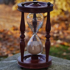 Könnte beinhalten: Eine Vintage-Sanduhr mit einem dunklen Holzrahmen und transparenten Glaskolben. Die Sanduhr enthält beigen Sand und wird von vier gedrechselten Holzbeinen getragen. Ober- und Unterseite sind kreisförmig, mit verschwommenem Herbstlaub im Hintergrund.