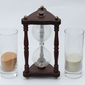 May include: A wooden hourglass with a dark brown frame and clear glass bulbs. Two clear glass tumblers, one filled with tan sand and the other with white sand, flank the hourglass. The hourglass is a decorative item.