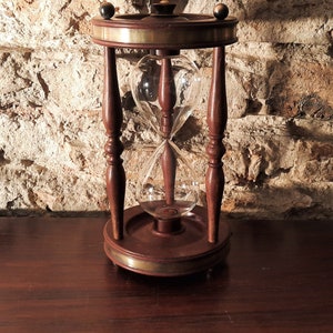 Könnte beinhalten: Eine Sanduhr aus Holz mit Messing-Oberteil und -Sockel. Die Sanduhr hat eine klare Glasbirne mit Sand im Inneren.