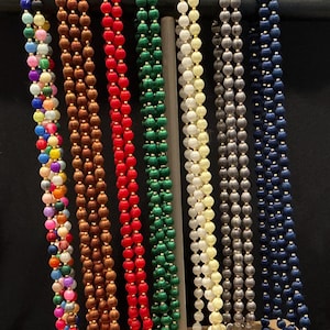 Puede incluir: Una colección de collares de cuentas en varios colores, incluyendo arcoíris, marrón, rojo, verde, crema, gris y azul marino. Cada collar presenta pequeñas cuentas redondas con detalles dorados, exhibidas verticalmente sobre un fondo oscuro.