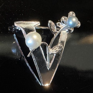 Peut inclure: Broche argentée en forme de lettre "V", ornée de deux perles et d'un motif floral. La broche est dotée d'une fermeture à épingle et est posée sur un fond noir. Les perles sont blanches et bleu clair.