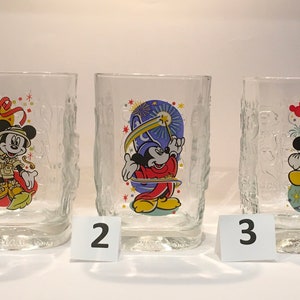 Op de afbeelding: Drie heldere drinkglazen met Mickey Mouse-ontwerpen. Elk glas heeft een andere kleurrijke illustratie van Mickey Mouse in verschillende poses. De glazen hebben een textuur, schorsachtig patroon aan de zijkanten.
