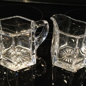 Puede incluir: Dos piezas de servicio de vidrio transparente facetado. Una es una jarra de crema con pico, y la otra es un azucarero con asa. Ambas tienen un diseño de estrella en la parte inferior y están sobre un fondo negro.