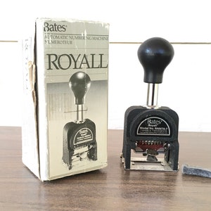 May include: A black Bates Royall automatic numbering machine with a black handle and seven wheels. The machine is in its original box, which is white with black text. The box says "Bates Automatic Numbering Machine Numeroteur Royall".